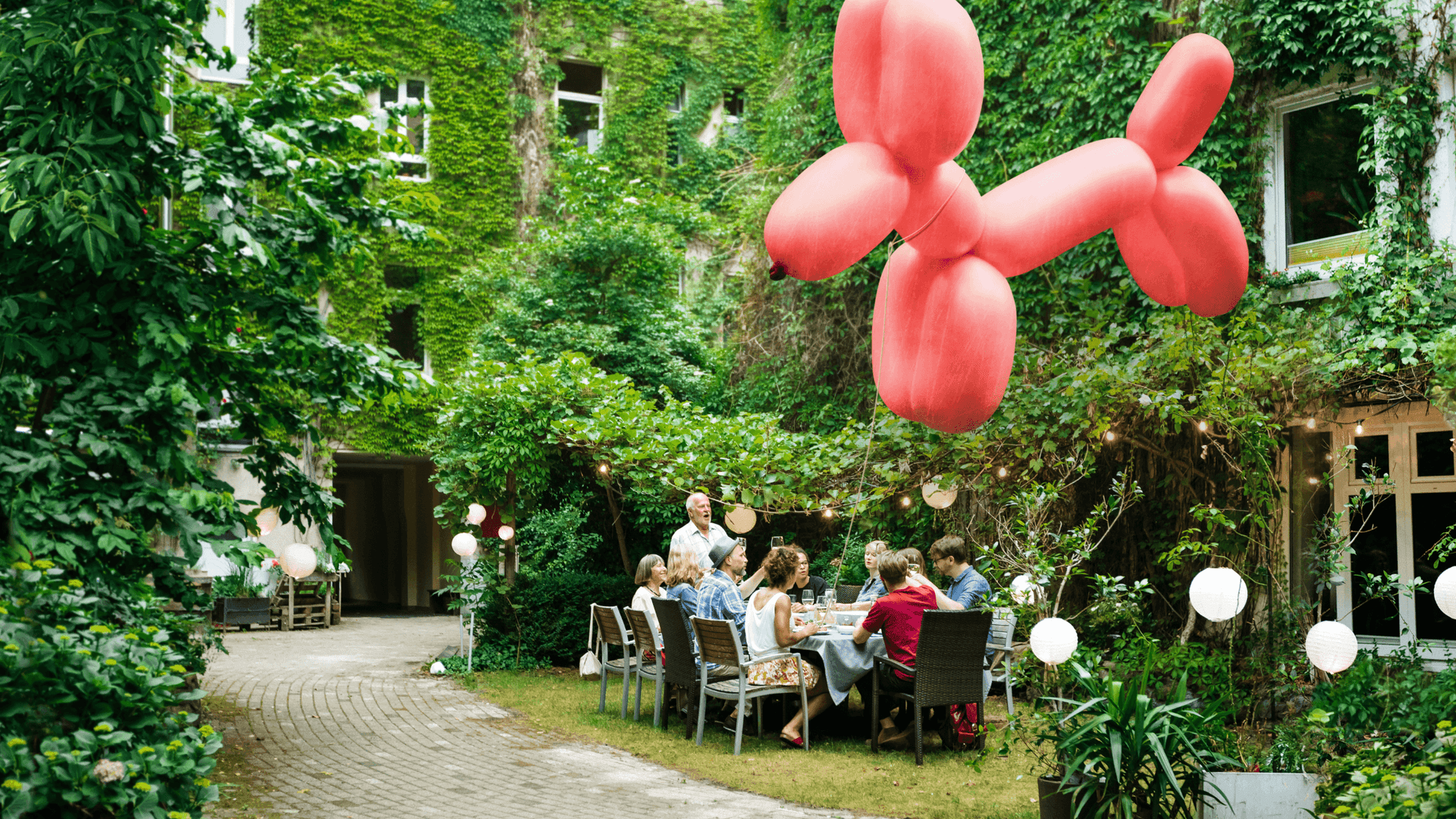 Ein pinkfarbener Ballonhund fliegt durch einen Hof, im Hintergrund sitzen Menschen beieinander.